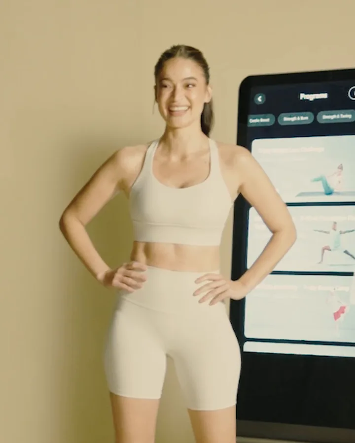 Anne Curtis smiles, hands on her hips, beside a ZYLO K1 screen showing workout programs.