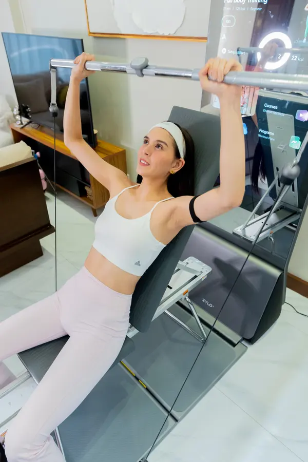 Ashley Ortega smiles, holding a cable bar overhead on the ZYLO bench, its lit screen behind her.
