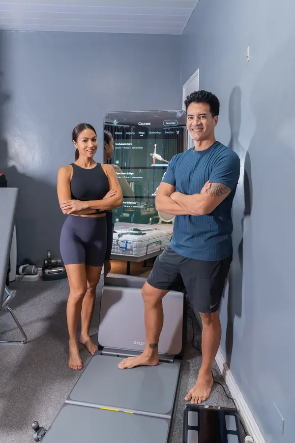 Aubrey and Troy stand with arms crossed beside a ZYLO K1 smart mirror, its screen lit.