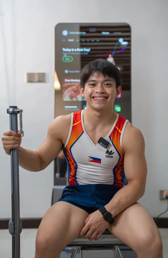 Carlos Yulo smiles, seated in front of a lit ZYLO K1 screen, holding an upright bar.