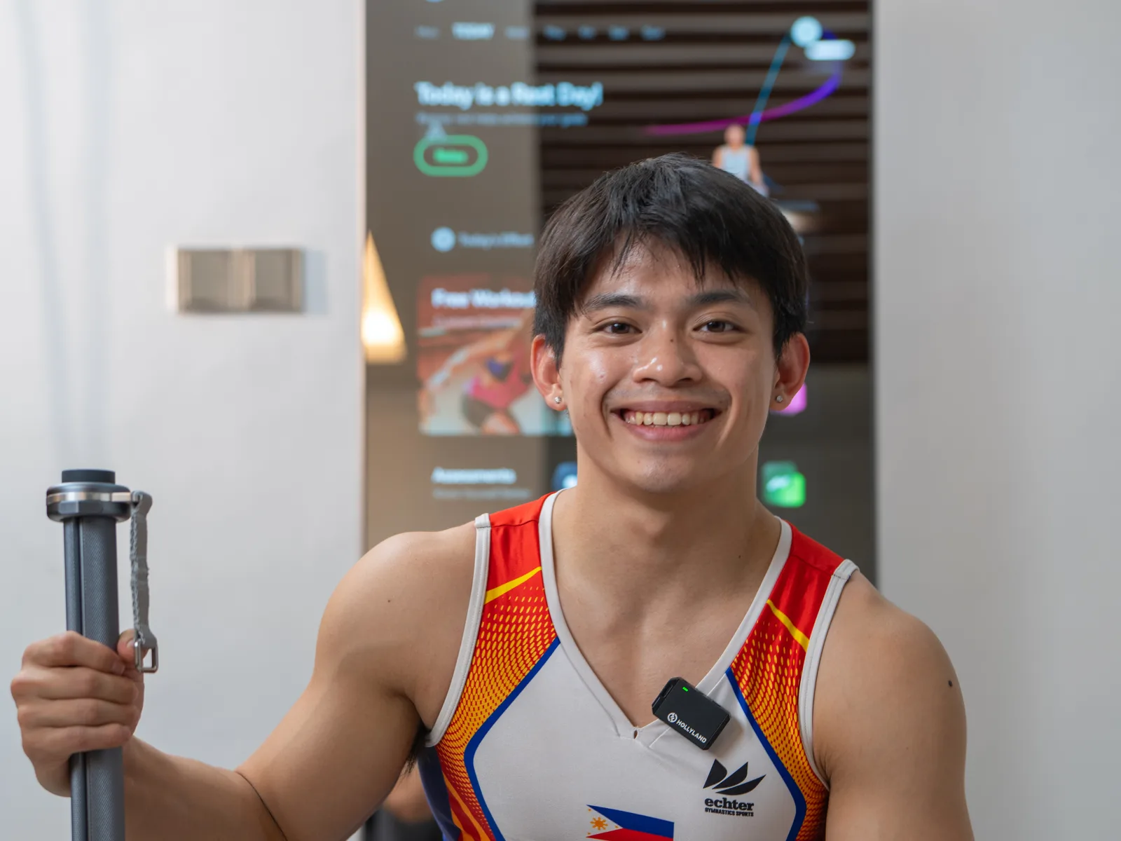 Carlos Yulo in his Philippines team singlet, smiling and holding a ZYLO cable handle in front of the lit K1 screen.