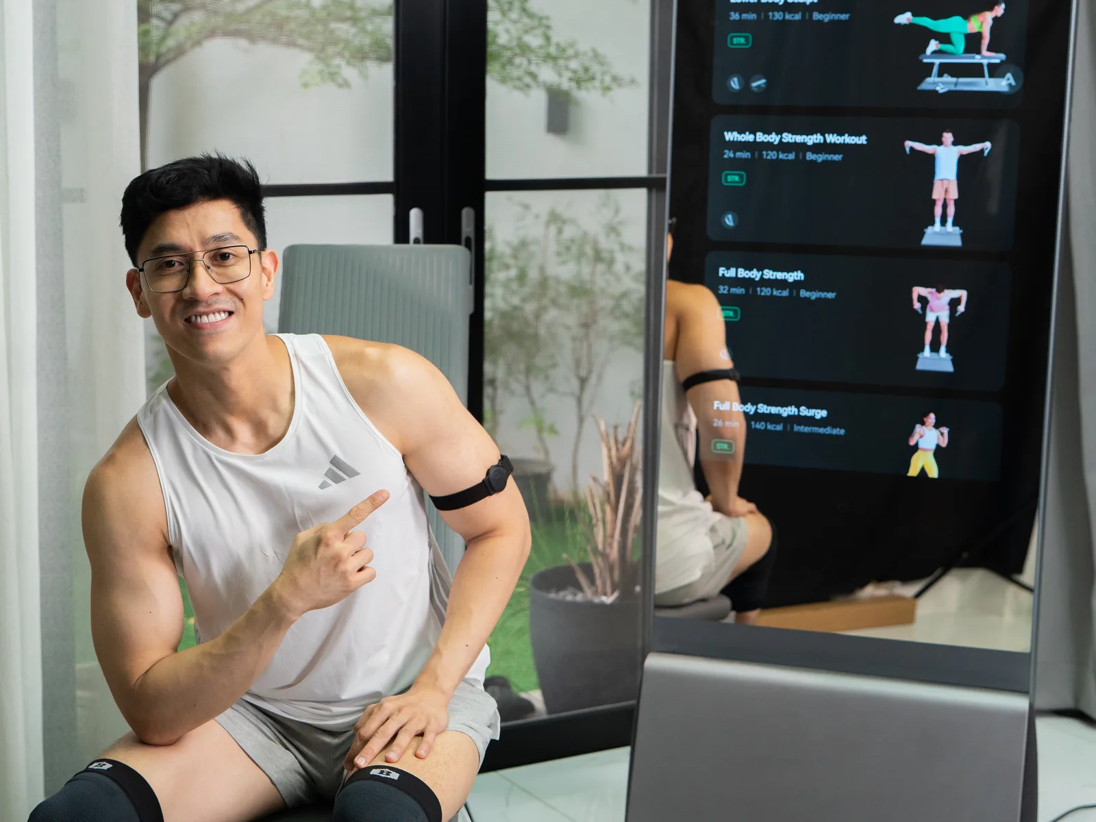 Doc Alvin seated beside his ZYLO K1 at home, gesturing towards it while the screen lists full-body strength workouts.